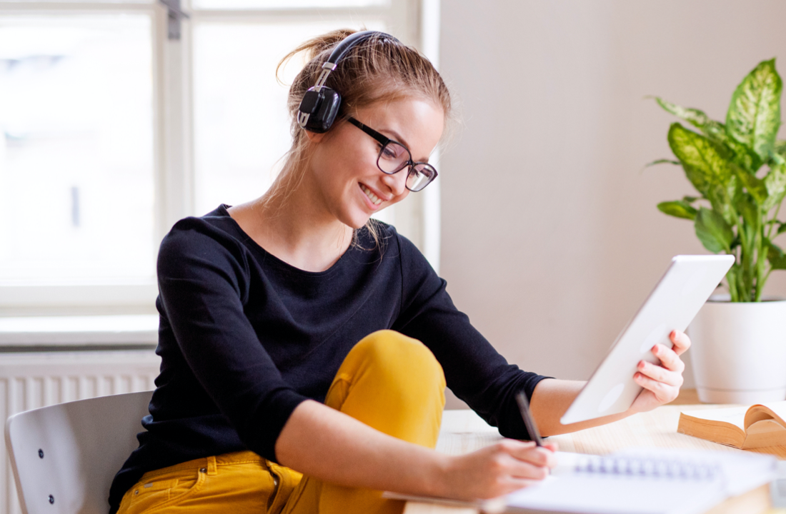 woman with headphones smiling at tablet and taking notes